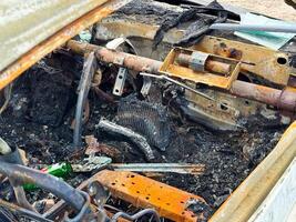 Detailed view of a burned car's dashboard area with rusted metal components, melted wiring, charred remains, and broken glass scattered on the scorched floor photo