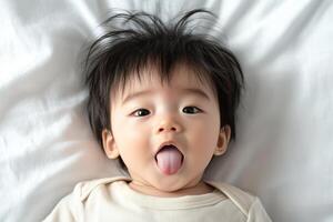 Cute Asian baby playfully sticking out tongue while lying on white bed photo
