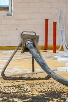 An active construction site showing a concrete pump hose on a tripod stand dispensing material onto a yellow surface, with white brick walls and red pipes. photo