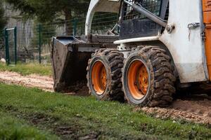 A compact skid steer loader with orange wheels and a front bucket rests on dirt and grass. Mud covered tires and a metal fence are visible nearby. photo