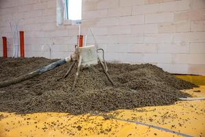 A construction site showing lightweight aggregate spread on a yellow vapor barrier. A mechanical tool disperses material near concrete walls and utility pipes. photo