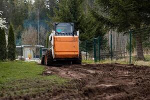 An orange and white skid steer loader works on a dirt path, leaving tire tracks. A green metal fence, tall evergreen trees, and a structure with a transparent roof are visible. photo