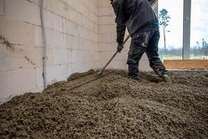 A worker uses a long handled tool to level wet concrete inside a building with white block walls. A large window reveals sparse trees and an open field. photo