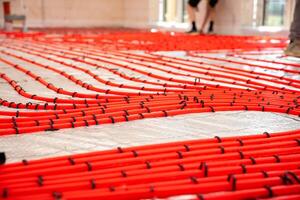 Bright red heating pipes arranged in a grid on reflective insulation, secured with black fasteners. Light walls, large windows, and workers' legs visible. photo