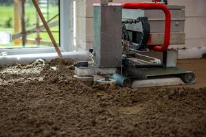 A concrete leveling machine with a red handle operates on wet material indoors. Natural light enters through a large window, revealing blurred green surroundings. photo