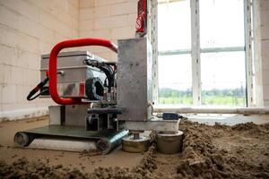 A construction site interior showing a machine with a red handle for underfloor heating pipe installation and Estrich concrete leveling on a lit concrete floor. photo