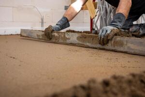A worker in gloves uses a straightedge tool to level wet concrete indoors. White walls and construction materials are visible in the background. photo