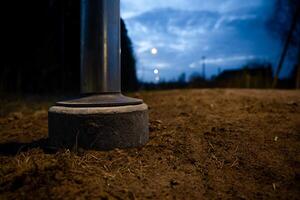 Low angle view of a metallic pole's base on a dirt pathway, with concrete foundation, faint streetlights, trees, and a partially clouded blue sky at dusk. photo