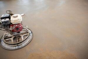 A power trowel machine with a red and white motor smooths a wet concrete surface. The circular blade contacts the floor, showing drying patches. photo