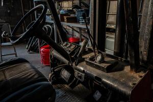 Weathered forklift interior showing steering wheel, control levers, and analog gauges. Industrial tools, hoses, and shelving visible in the setting. photo
