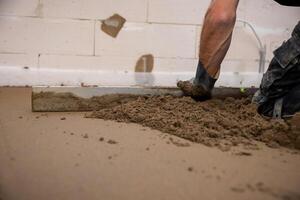 A worker uses a leveling tool to smooth Estrich concrete over underfloor heating pipes. Partially finished walls and exposed material are visible indoors. photo