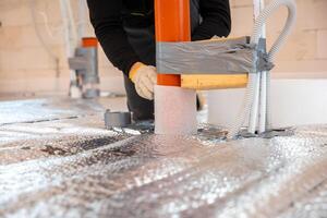 A worker secures red underfloor heating pipes with duct tape and foam insulation on reflective foil. Tools, materials, and unfinished walls are visible. photo