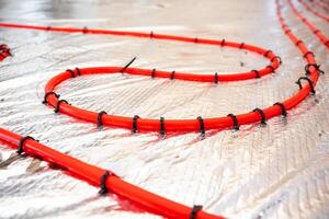 Bright red heating pipes are coiled and secured with black fasteners on a reflective foil insulation layer, highlighting the installation process. photo