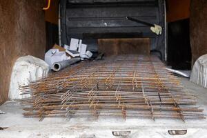 Stack of rusted metal mesh sheets inside a vehicle cargo area with wooden paneling, pipes, and boxes, suggesting preparation for a construction project. photo