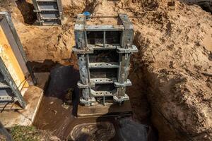Metal framework structure partially embedded in sand and soil, with bolts and clamps visible. A pool of water near the base shows excavation activity. photo