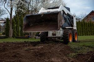 A skid steer loader with orange wheels rests on freshly dug soil near trees with white blossoms, trimmed hedges, and a house with a sloped roof. photo