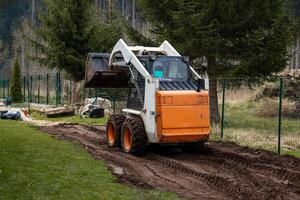 A compact orange and white skid steer loader operates on a dirt path, leaving tire tracks. Surrounded by a wire fence, evergreen trees, logs, and bags. photo