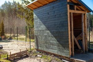 A dark green wooden shed with a slanted roof stands partially built on dirt near a fenced area, with sparse trees and a forested area in the background. photo
