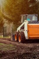 An orange and white skid steer loader rests on a muddy path with its bucket down, surrounded by a wire fence and tall evergreen trees in sunlight. photo