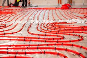 Bright red underfloor heating pipes arranged in coiled patterns on a reflective surface, with workers in high visibility clothing in the background. photo
