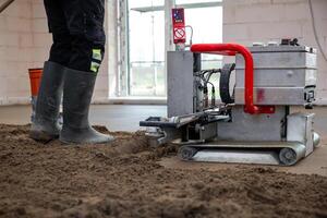 A construction worker operates a leveling machine with a red handle on concrete in an unfinished interior with white brick walls and large windows. photo