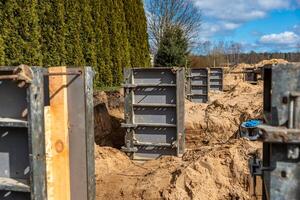Metal framework panels stand on sandy soil at a construction site. A tall hedge borders one side, with leafless trees and sparse vegetation visible. photo