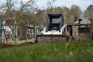 A white skid steer loader with its bucket lowered sits on grass, surrounded by sparse trees, a fenced area, a shed, and a house in a rural setting. photo