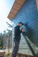 A man in dark clothing works with a yellow power drill on a slanted wooden structure. The setting includes a fenced area and trees under bright sunlight. photo