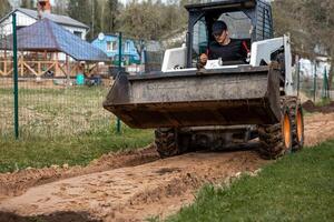 A man uses a skid steer loader with a front bucket to level a dirt path. Green wire fence and residential houses with pitched roofs are visible nearby. photo