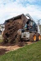 A skid steer loader with orange wheels dumps soil onto the ground, operated by a worker. Industrial buildings and trees are visible in the surroundings. photo
