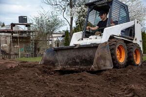 A man uses a skid steer loader with orange wheels to move soil. The area includes trees, some in bloom, a shed, fencing, and natural surroundings. photo