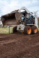 A skid steer loader with orange wheels raises a bucket filled with soil. The scene shows freshly tilled ground, a wire fence, and distant structures. photo