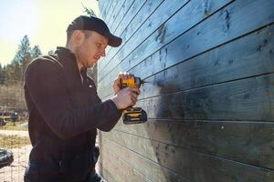 A man in a black cap and jacket operates a yellow and black power drill on a dark wooden wall. Trees and a clear blue sky are visible in the background. photo