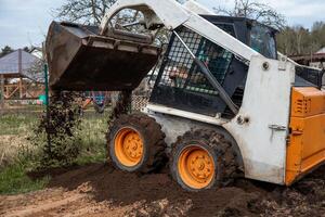 A white and orange skid steer loader with raised bucket dumps soil on uneven ground. Nearby are a fenced area, playground equipment, and sparse trees. photo