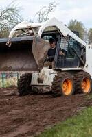 A skid steer loader with orange wheels carries soil in its raised bucket. A person operates the machine in a grassy field with sparse trees nearby. photo