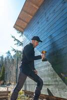 A man in dark clothing and a black cap operates a yellow power drill on a wooden structure with a slanted roof. Tall trees and a metal fence are visible. photo