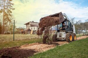 A skid steer loader with orange wheels dumps soil on a pathway near a wooden house with a slanted roof, surrounded by fields and sparse trees. photo