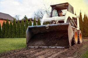A white skid steer loader with orange wheels levels soil on a dirt path. A person operates it near tall evergreen trees and a slanted roof structure. photo