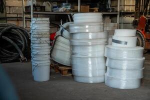 Concrete floor workspace featuring stacked white buckets, foam rolls, coiled hoses, tools, shelves with supplies, and a partially visible forklift. photo