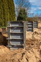 Metal framework panels rest in sandy soil at a construction site, surrounded by evergreen trees and a leafless deciduous tree, with visible trenches. photo