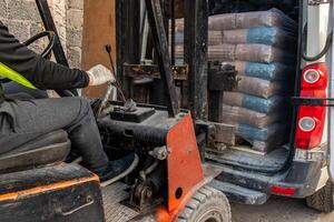 A worker operates an orange and black forklift to load plastic wrapped bags into a van. A stone wall and part of a building are visible nearby. photo