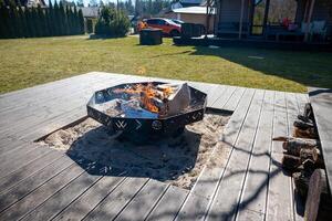 A fire pit with decorative patterns burns on a wooden deck, surrounded by a grassy yard. A modern house, porch, and red car are visible in the background. photo