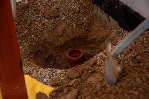 A construction site featuring a partially dug hole with a red brown pipe in the soil, loose dirt, gravel, a shovel, and a concrete wall with black waterproofing. photo