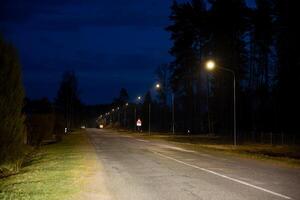A road extends under a deep blue evening sky, bordered by trees and grass. Streetlights illuminate the pavement, with a triangular traffic sign visible. photo
