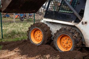 A compact skid steer loader with orange wheels moves soil on uneven ground. Rugged tires and a fenced area with playground equipment are visible. photo