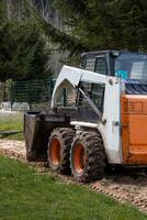 A white and orange skid steer loader with a bucket attachment sits on dirt and grass. A green metal fence and tall evergreen trees are in the background. photo
