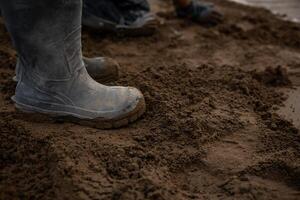 Muddy boots stand on damp, uneven soil with additional footwear in the background. The ground appears freshly disturbed, suggesting outdoor work activity. photo