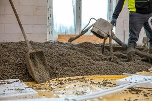Construction workers pour and level concrete over yellow plastic sheeting and insulation panels, with tools and bare trees visible through window openings. photo