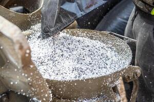 Close up of a metallic container as white granules are poured from a black bag, with machinery and a worker partially visible on the side. photo
