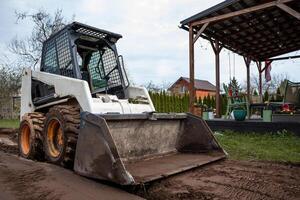 A skid steer loader with muddy tires rests on a dirt path near a wooden pergola with string lights, outdoor furniture, a red house, and bare trees. photo